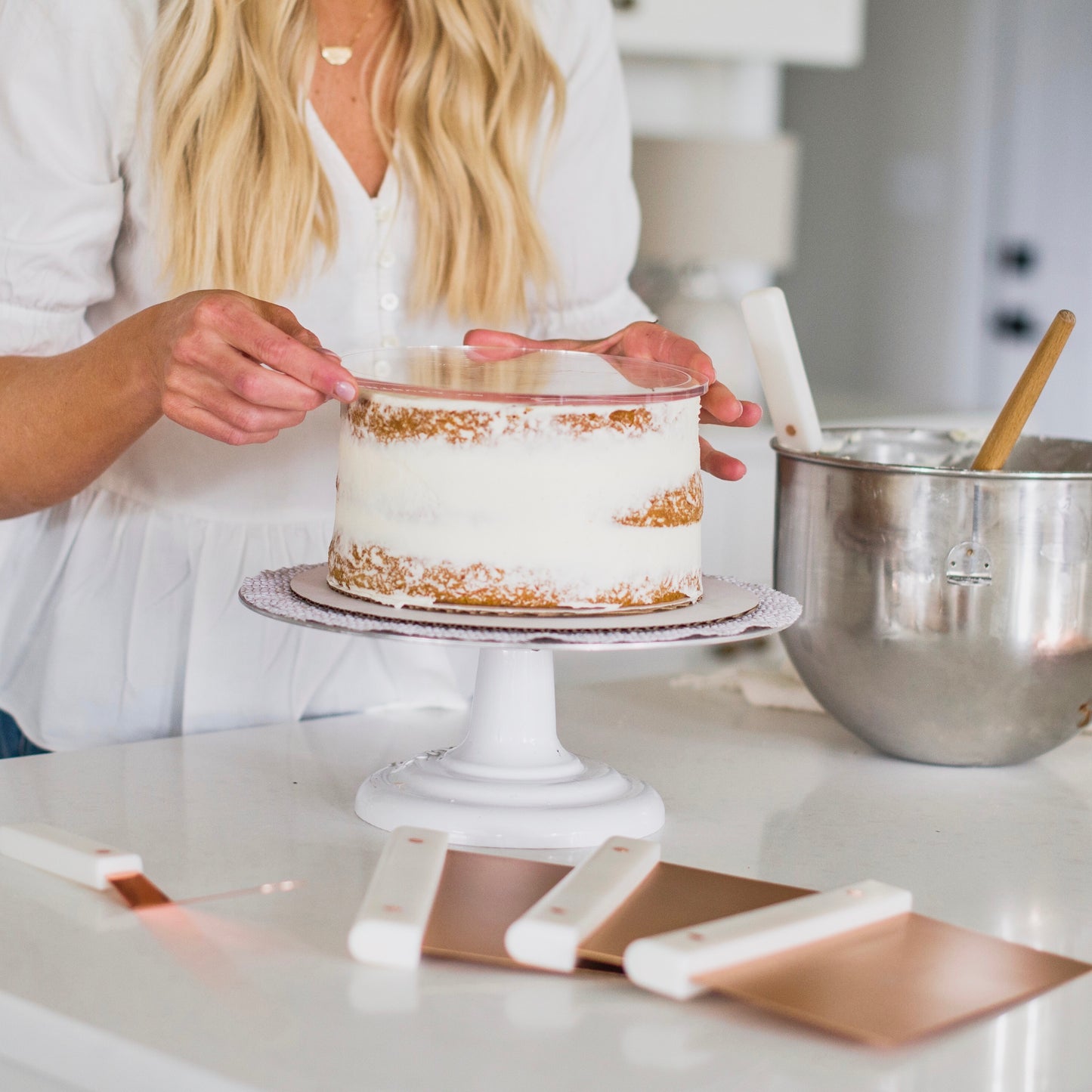 Courtney in a white top putting her acrylic disk on top of a cake to start icing, surrounded by her scrapers and spatula