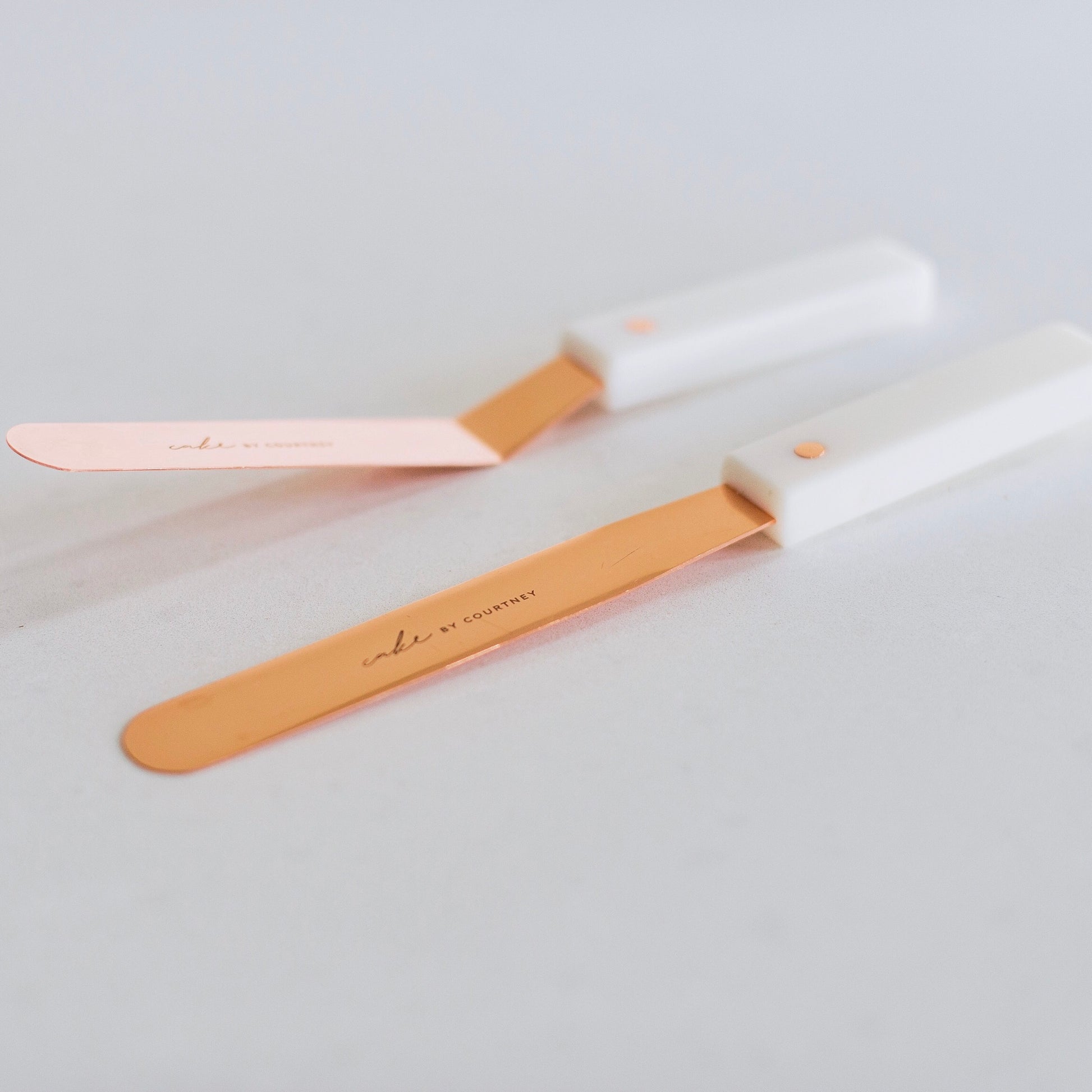 Close up of Courtney's two rose gold icing spatulas against a white countertop. One is angled and one is completely flat.