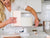 Courtney wearing a white blouse in her kitchen, using her flat scraper to smooth out the sides of her cake.