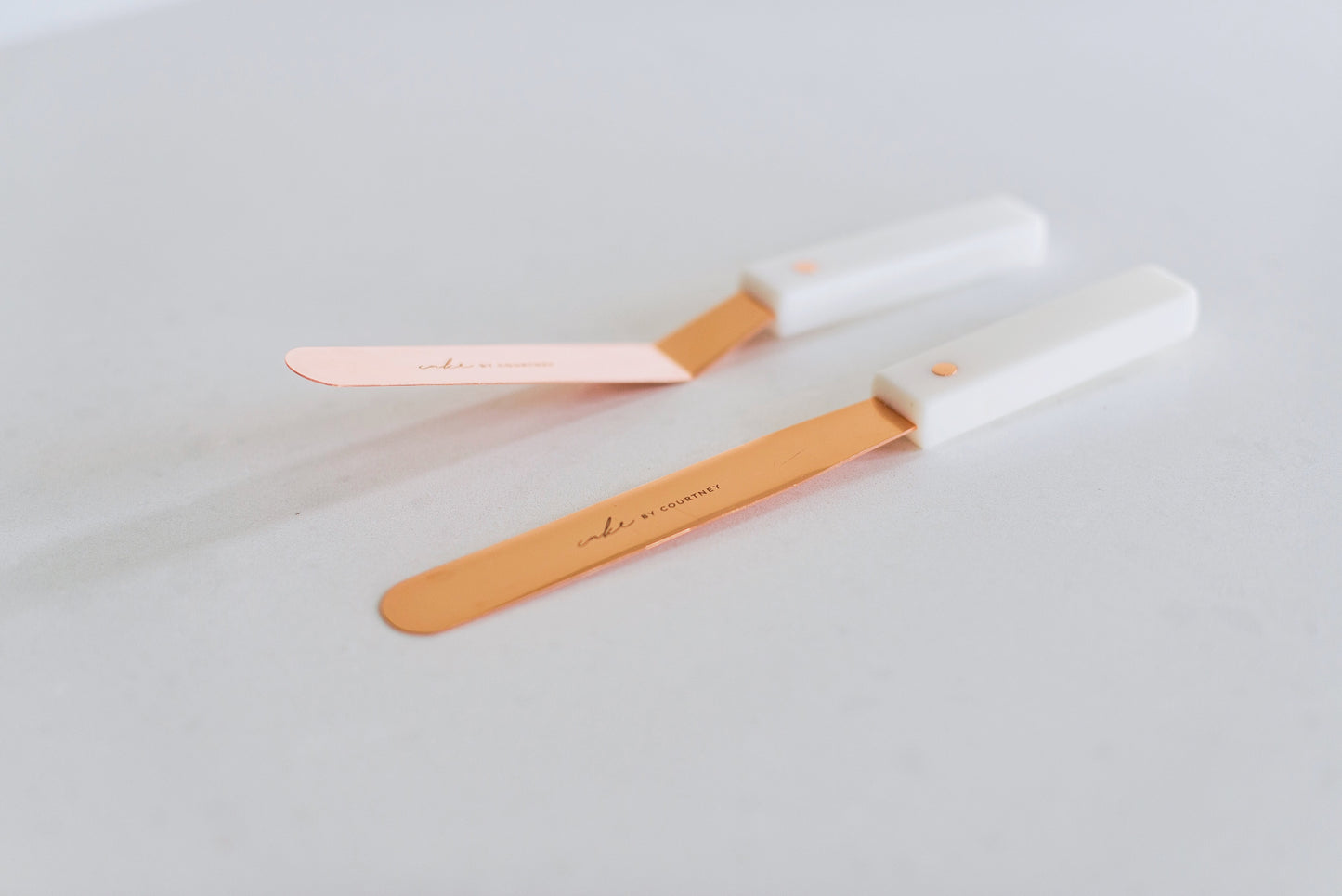 Close up of Courtney's two rose gold icing spatulas against a white countertop. One is angled and one is completely flat.