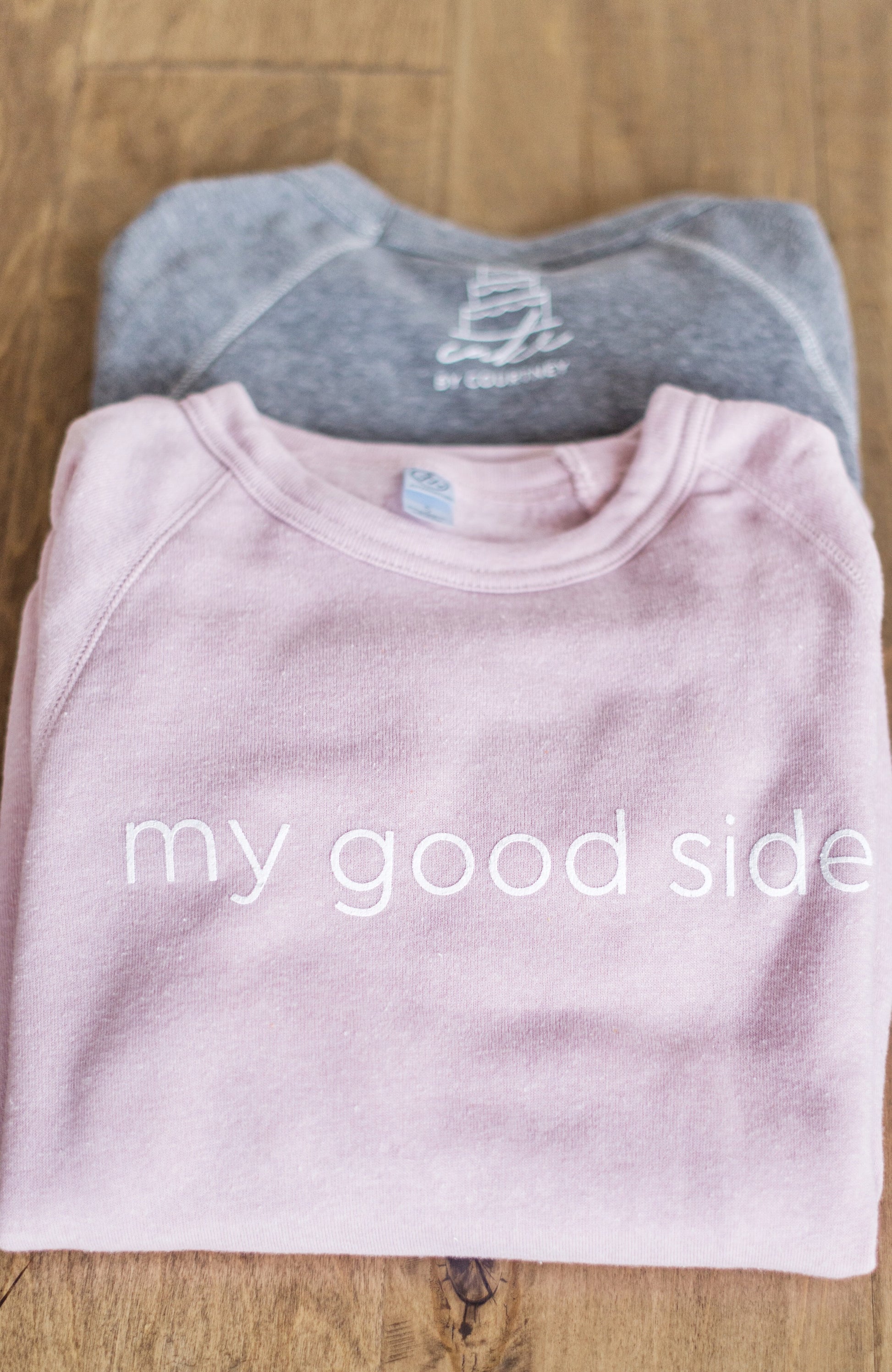 Lay flat image of both the grey and pink 'my good side' sweatshirt folded on a wood surface