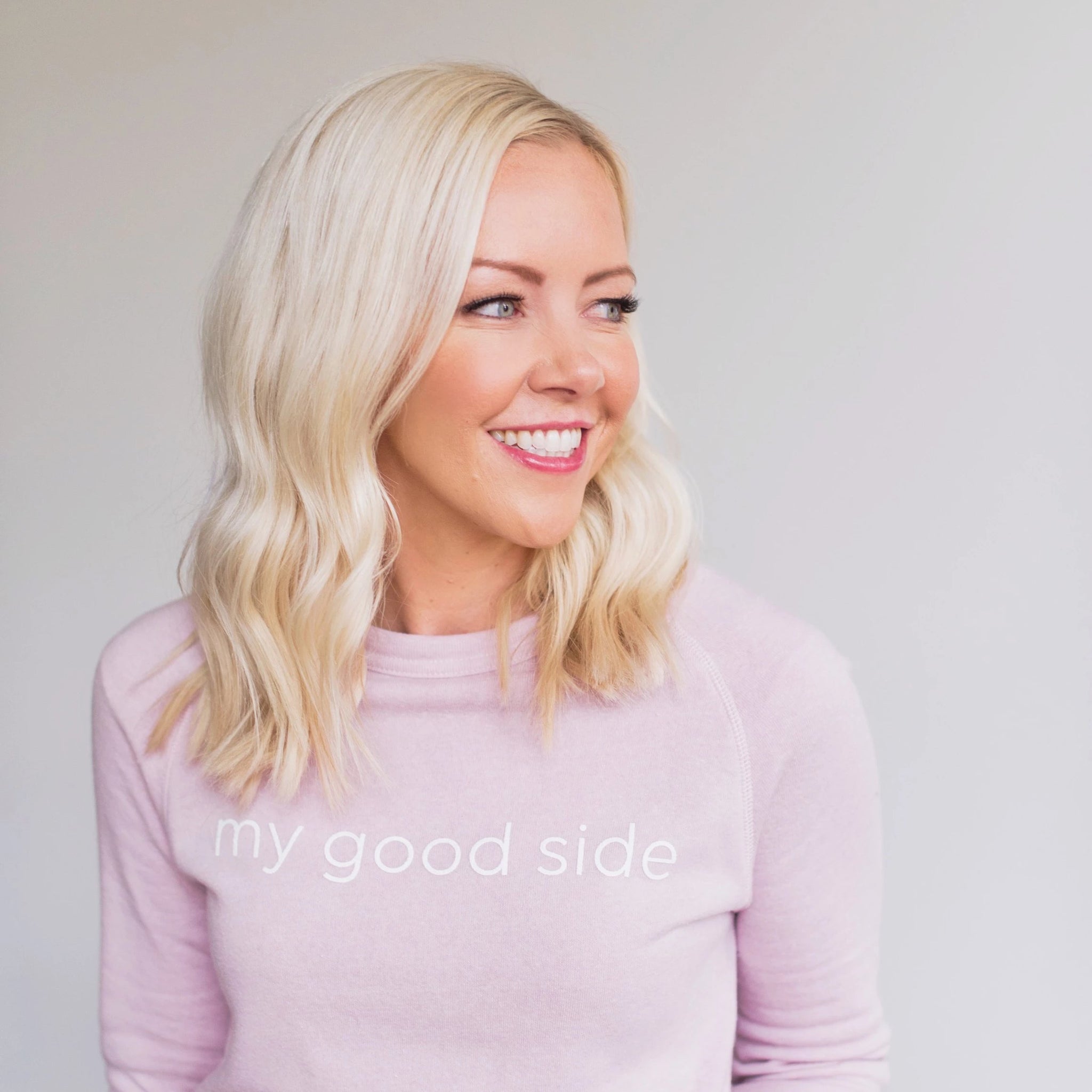Courtney smiling to the side, wearing her light pink sweatshirt that says the words 'my good side' that can be purchased on the website